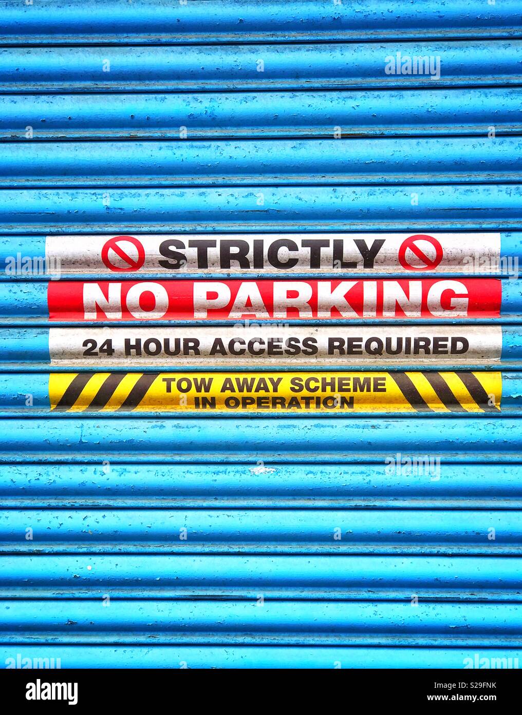 A blue roller shutter door cover d in warning signs that advise No Parking is permitted in this area. Constant access is required to this building. Your vehicle will be removed. You have been warned! - Smartphone Captured Stock Image