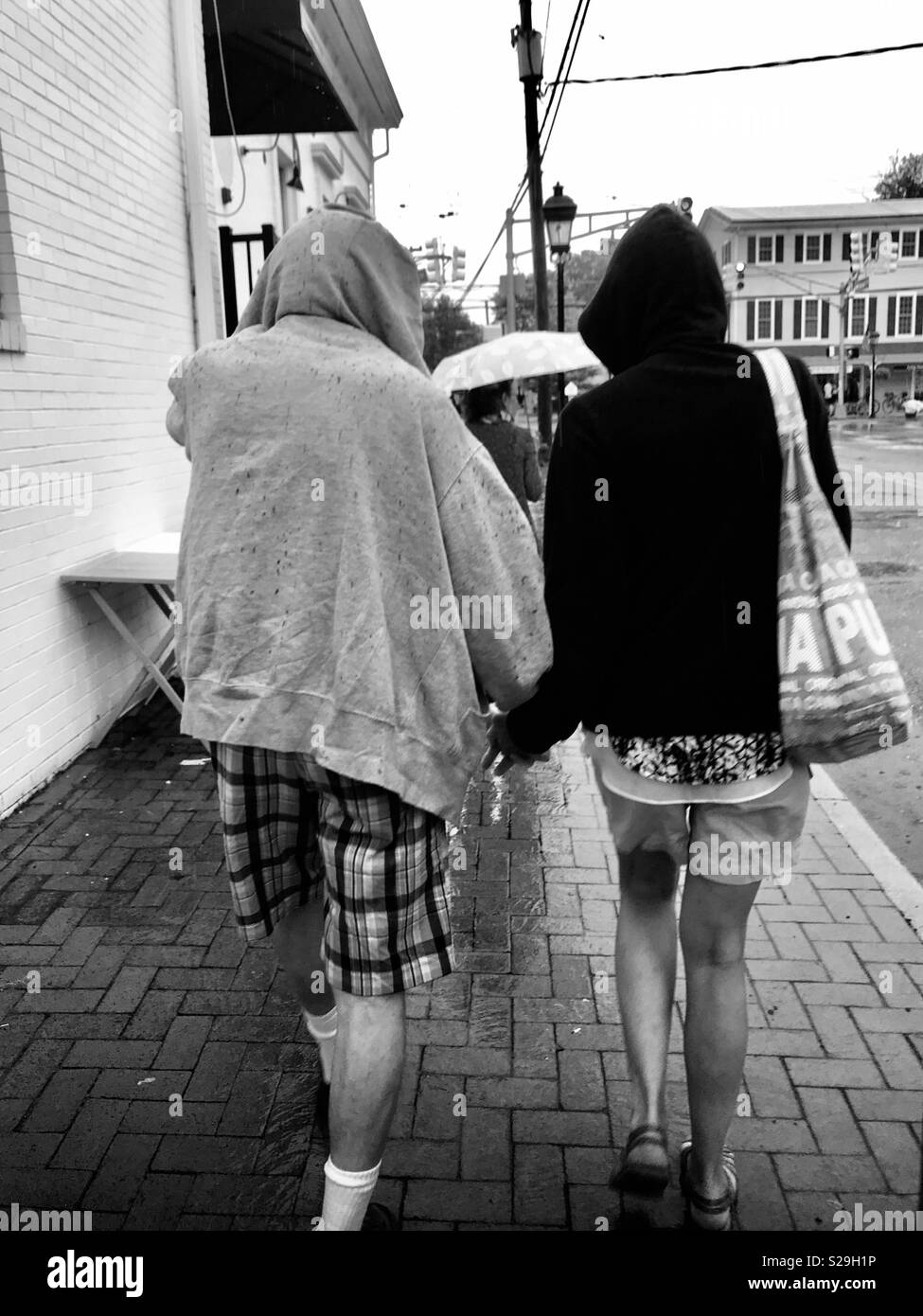View of the backs of an older couple wearing hoods walking hand in hand in the rain. They are wearing shorts and sweatshirts. Black and white photograph. Walking stay from the camera. Stock Photo