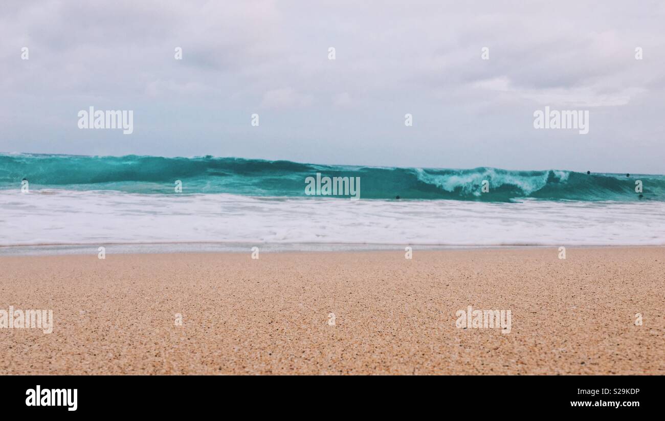 Looking at large waves from sandy beach on an overcast day with surfers in the distance. - Smartphone Captured Stock Image