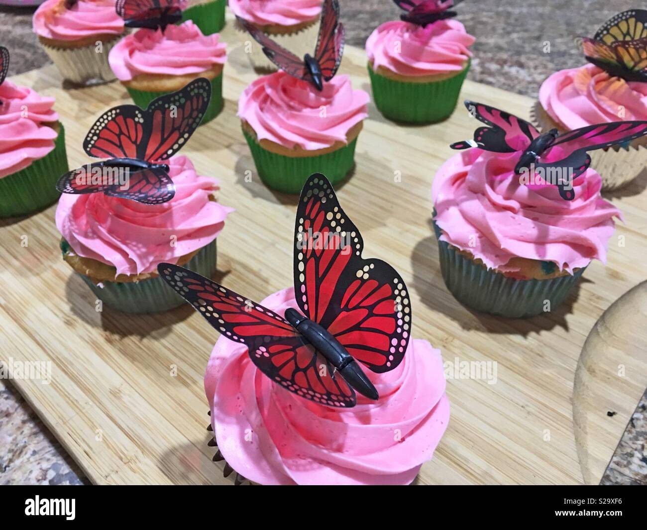 Cupcakes with hot pink icing and colorful red and black butterflies on top displayed on a wooden cutting board. - Smartphone Captured Stock Image