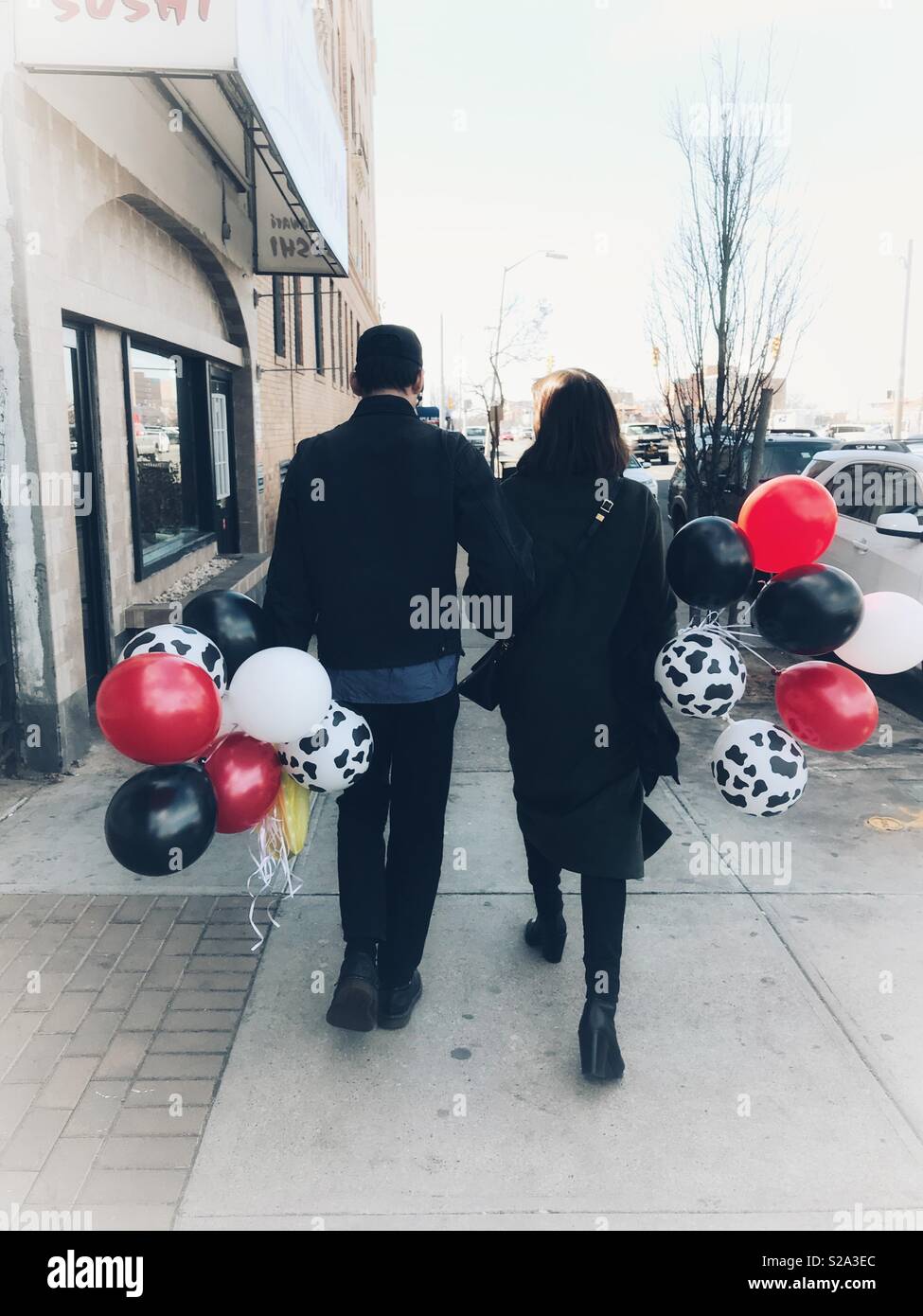 Man and woman in long coat walking away down the sidewalk arm in arm with each holding a bunch of colorful balloons. Background is blown out. Stock Photo