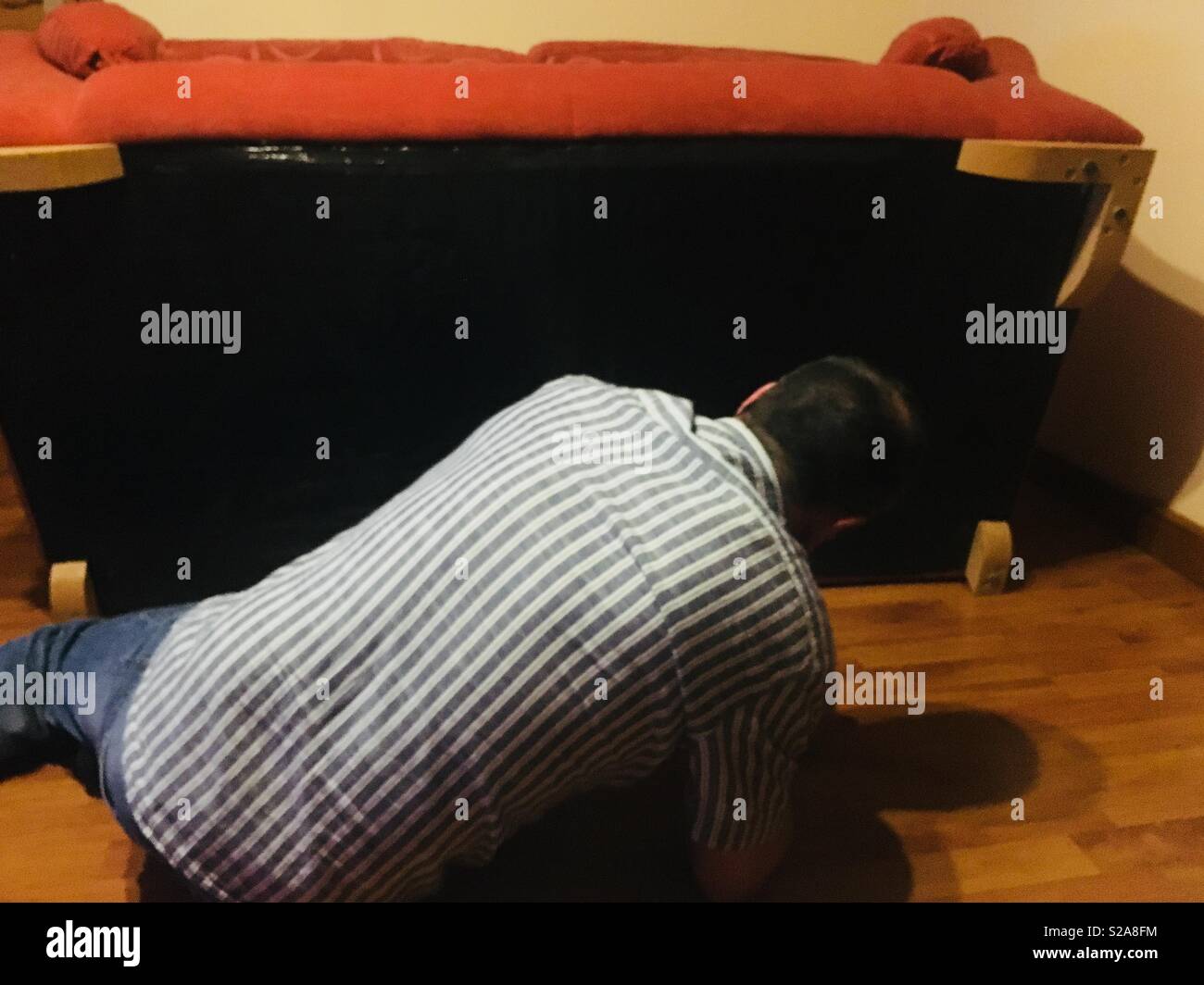 Man repairing the underneath of a terracotta coloured sofa whilst lying on a laminate floor Stock Photo