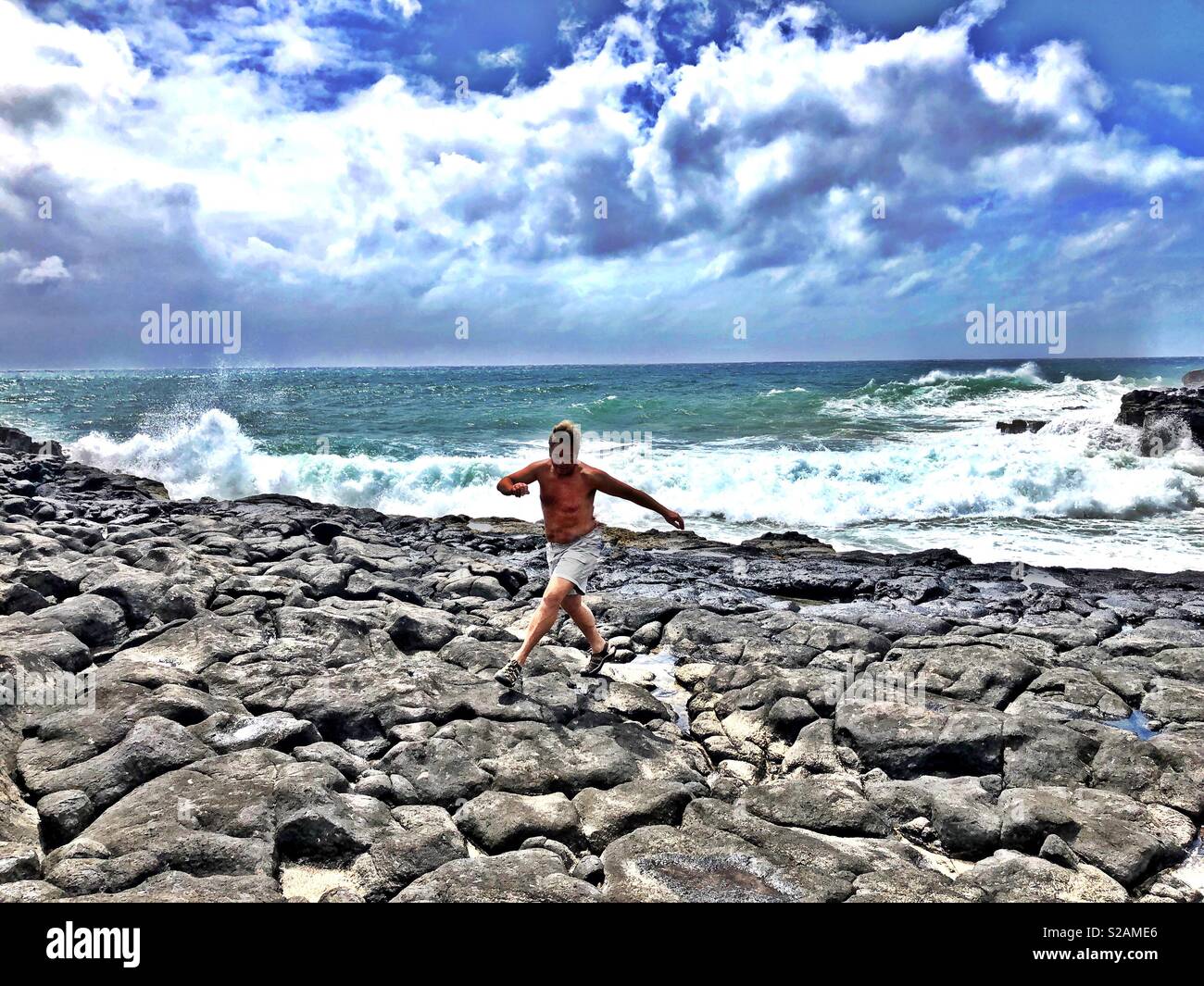 Athletic blond man running across lava rock on the rough southeastern shore of Kauai with waves crashing in the background as ominous clouds press against a tropical azure sky - Smartphone Captured Stock Image