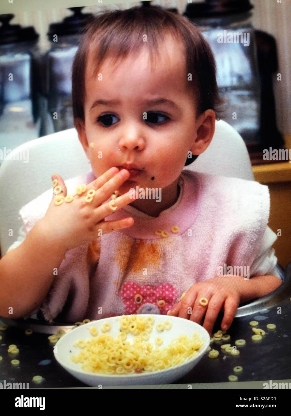An infant girl in a high chair messily eating noodles - Smartphone Captured Stock Image
