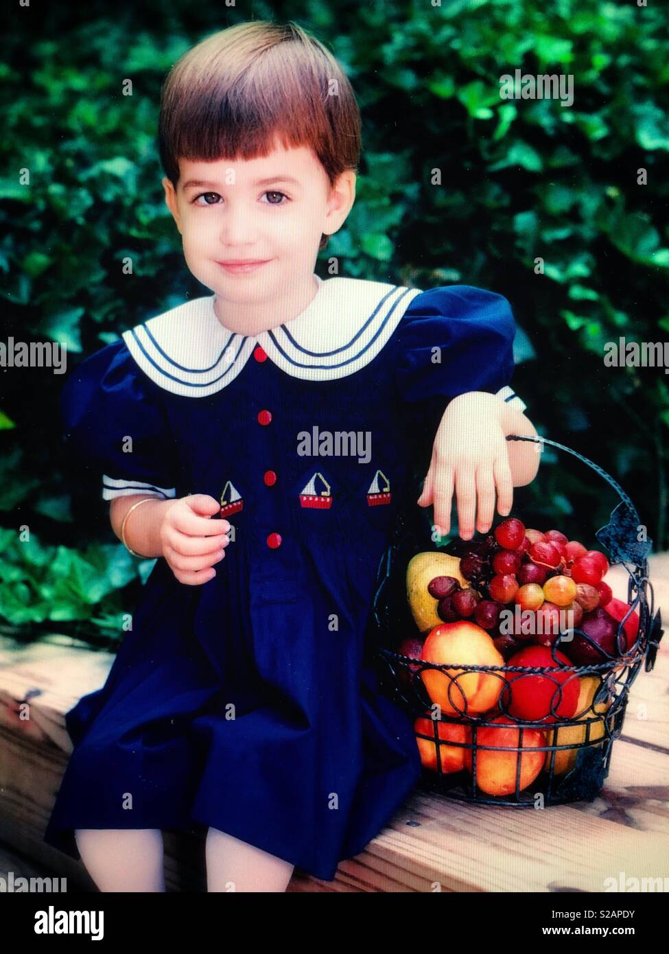 A toddler girl posing and smiling with a basket of fruit - Smartphone Captured Stock Image