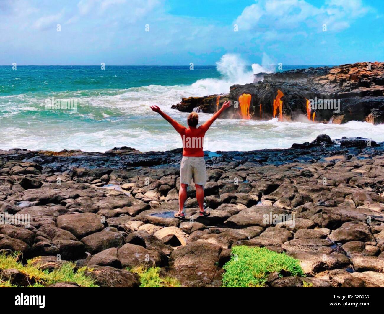 Tanned man arms outstretched embracing fresh hot lava from Goddess Pele flowing into the tumultuous ocean waves below on the island of Hawaii - Smartphone Captured Stock Image