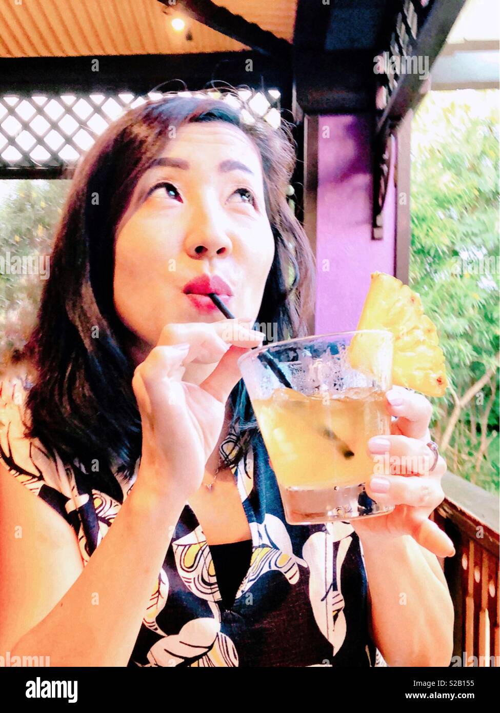 Girl with mai tai during happy hour at a tropical island beachcomber tiki bar coquettishly daydreams with smiling eyes - Smartphone Captured Stock Image