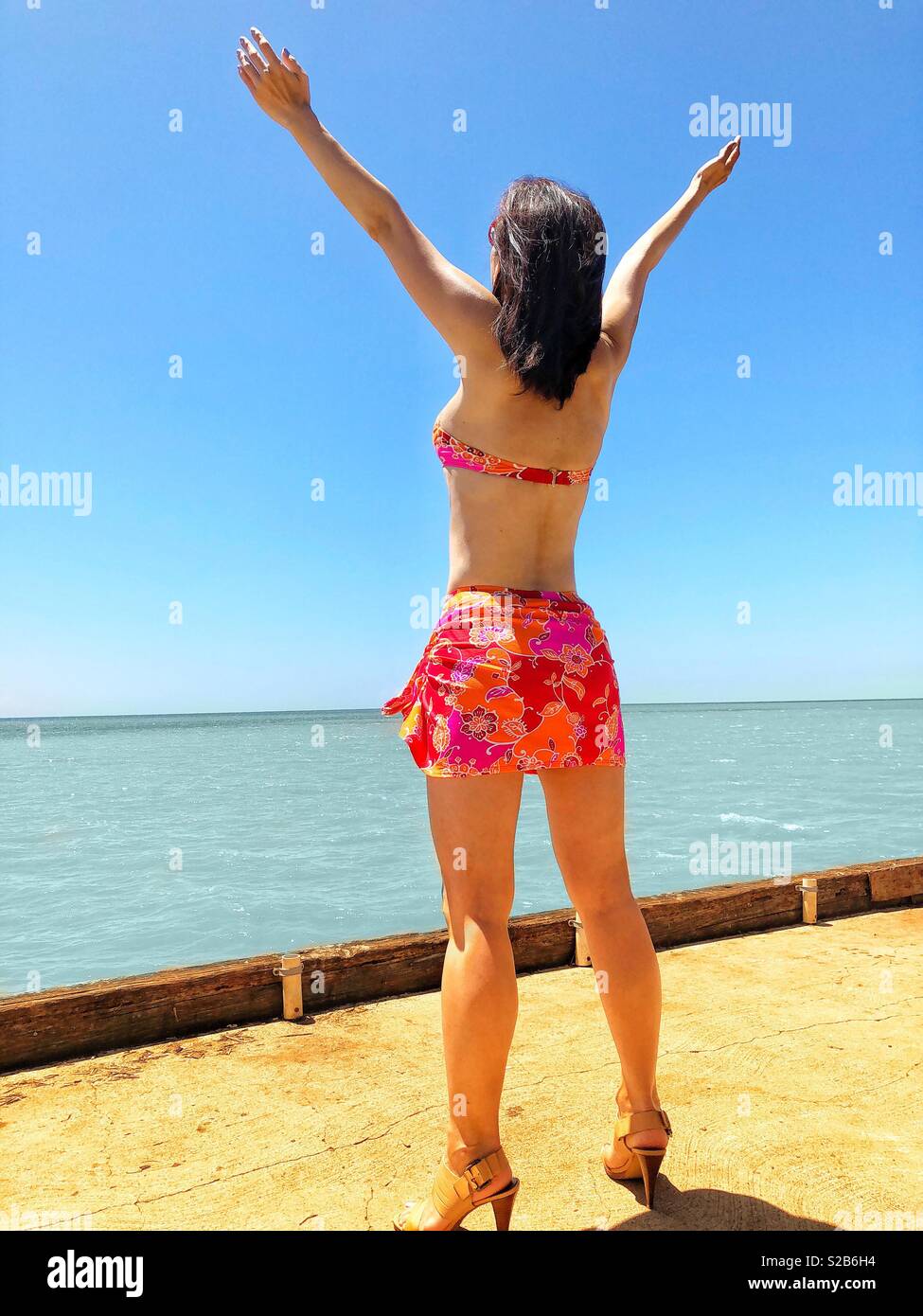 Young brunette woman in floral pink bikini and paleo skirt embraces the tropical Hawaiian island shore at the end of a pier on a bright sunny day in paradise Stock Photo
