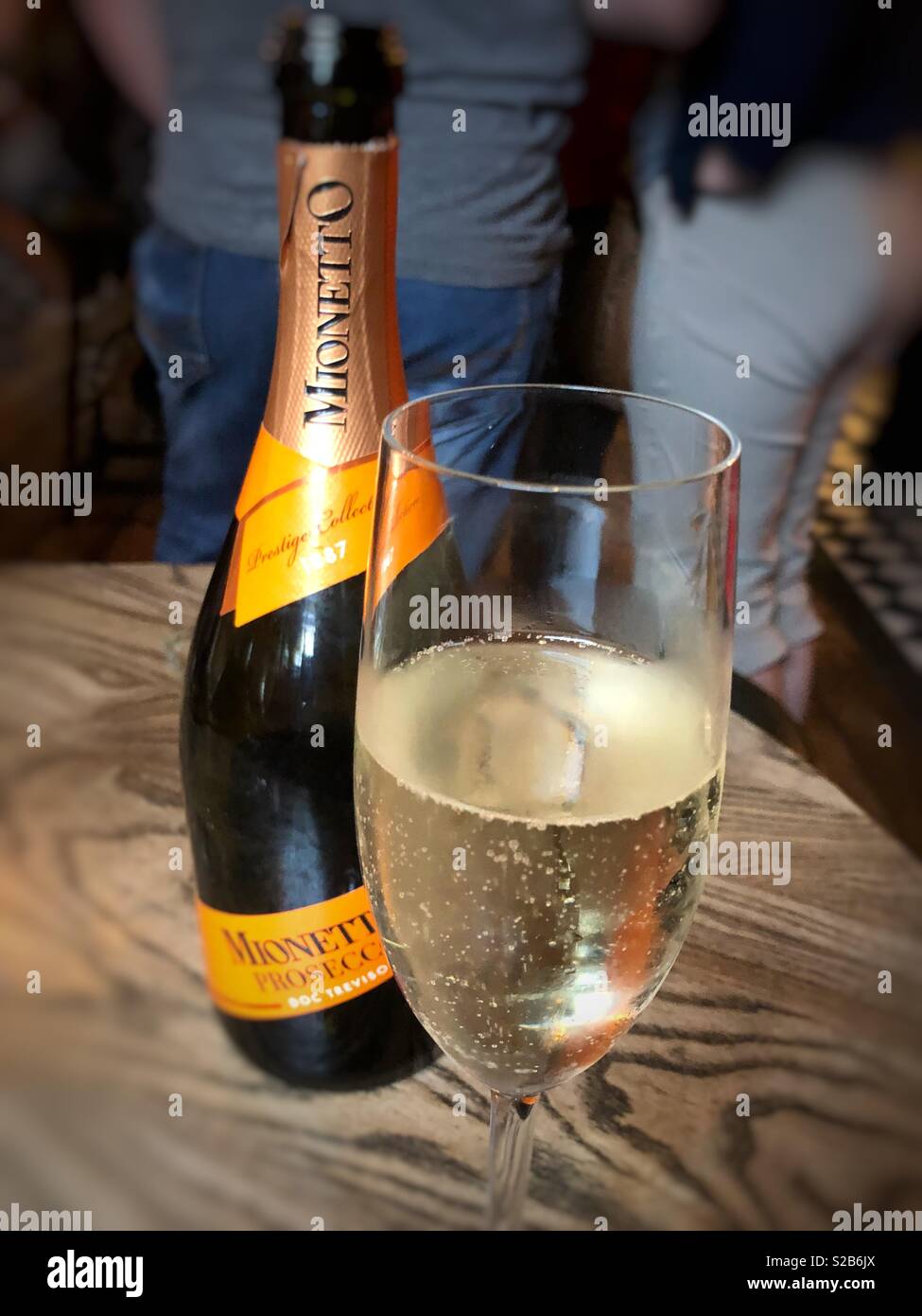 A Bottle of prosecco with a half full glass next to it in a pub with a man and a woman blurred in the background Stock Photo
