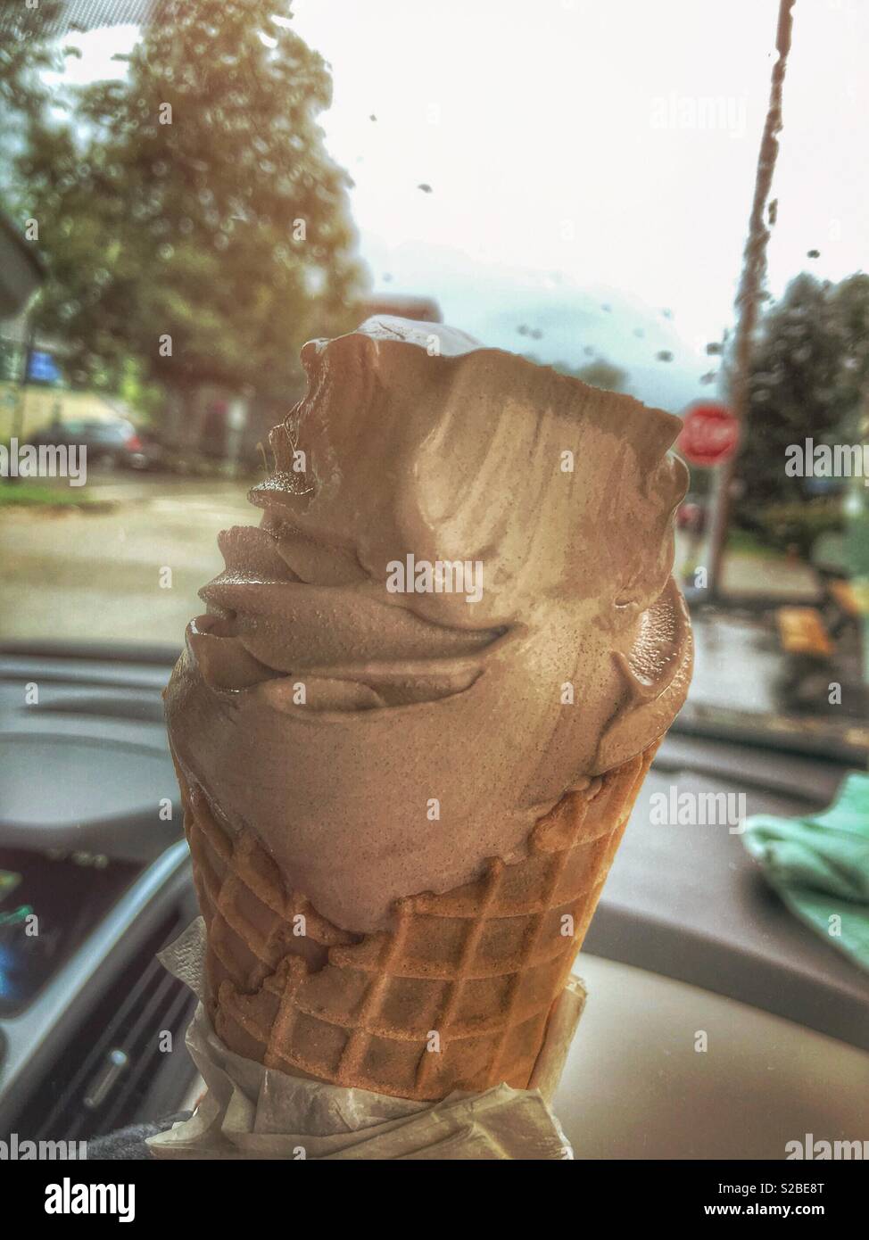 Chocolate soft serve ice cream in a waffle cone.  It is being enjoyed on a rainy day in a car. Rain drops are on the windshield, and a stop sign can be seen in the distance. Stock Photo