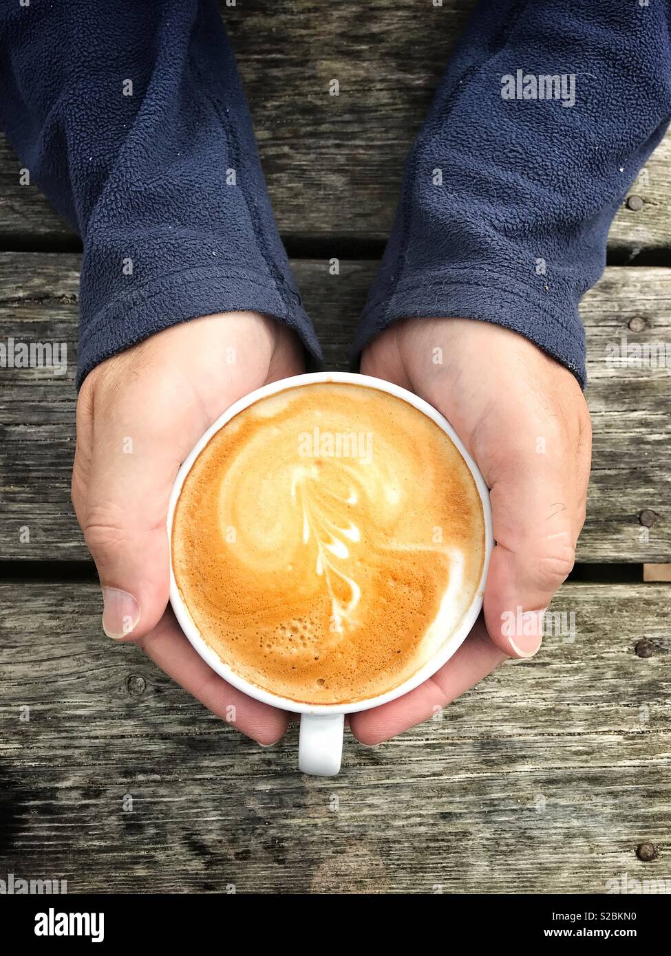 Looking down from above onto a a pair of male hands cupping a hot cup of cappuccino coffee on a wooden table - Smartphone Captured Stock Image