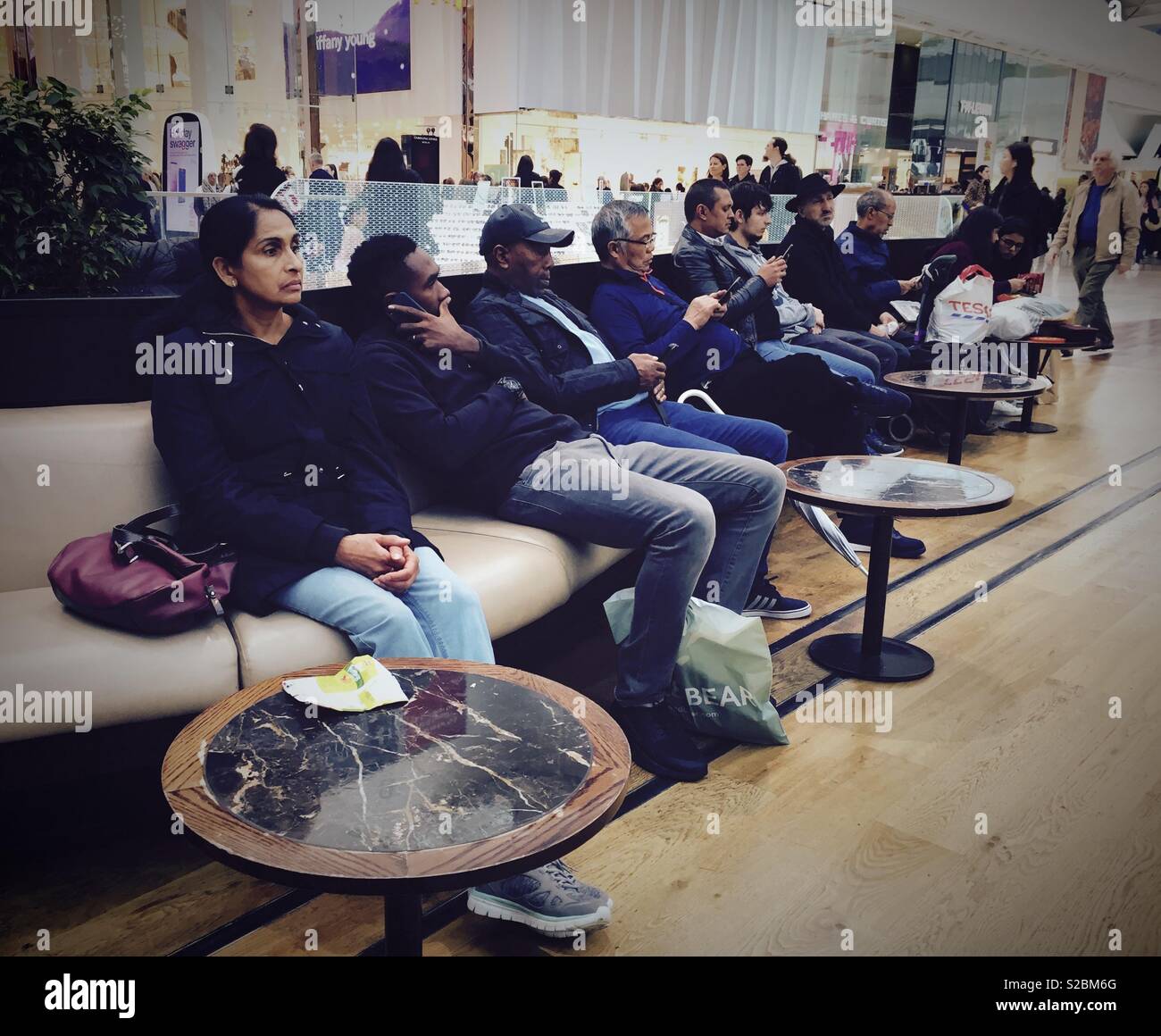 People sit, rest and check their phones on seating in a shopping centre. - Smartphone Captured Stock Image