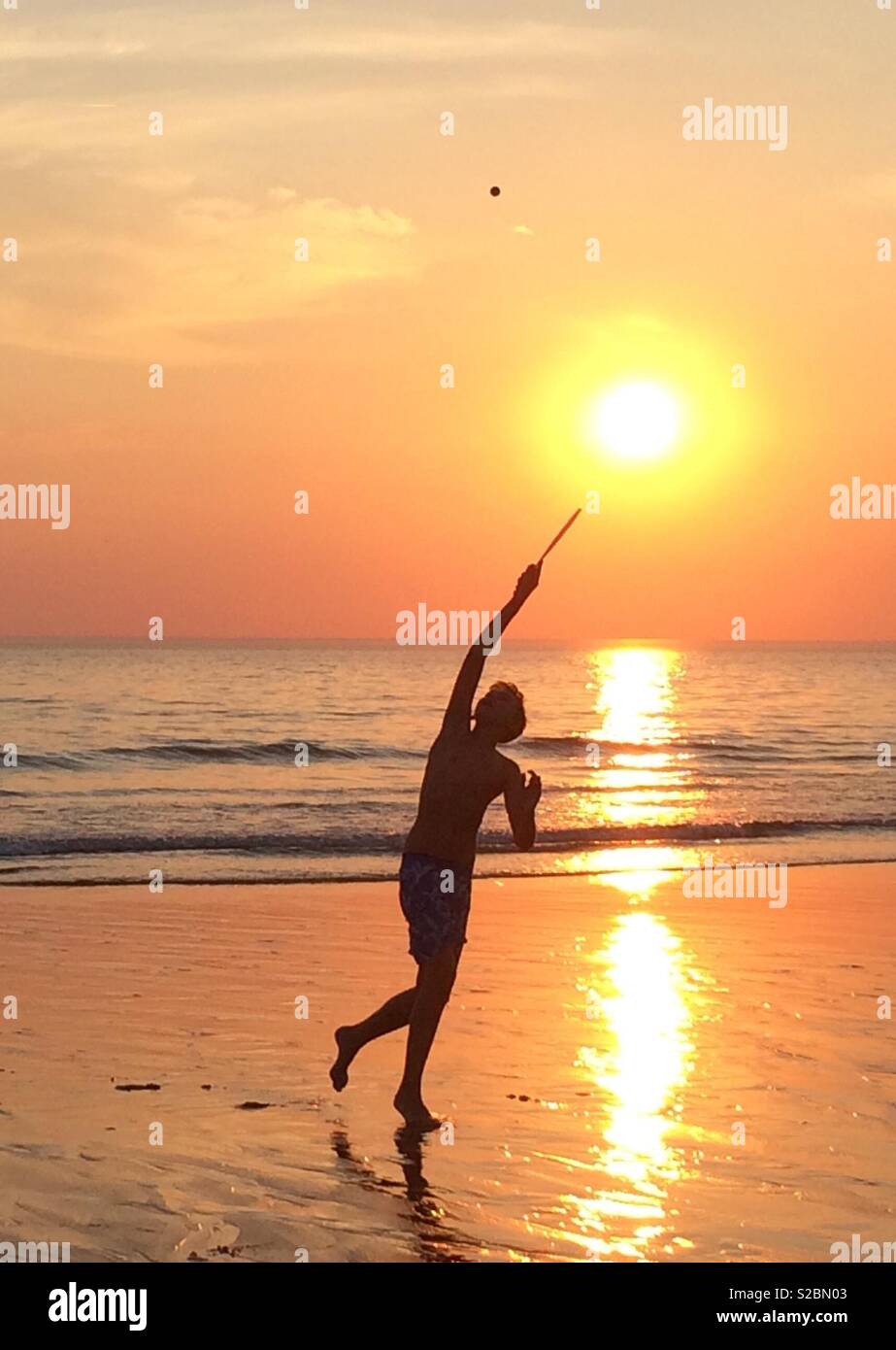 Silhouette of a boy playing bat and ball on a beautiful French beach, early evening as the sun was setting, reflected on the golden shining sand. Stock Photo