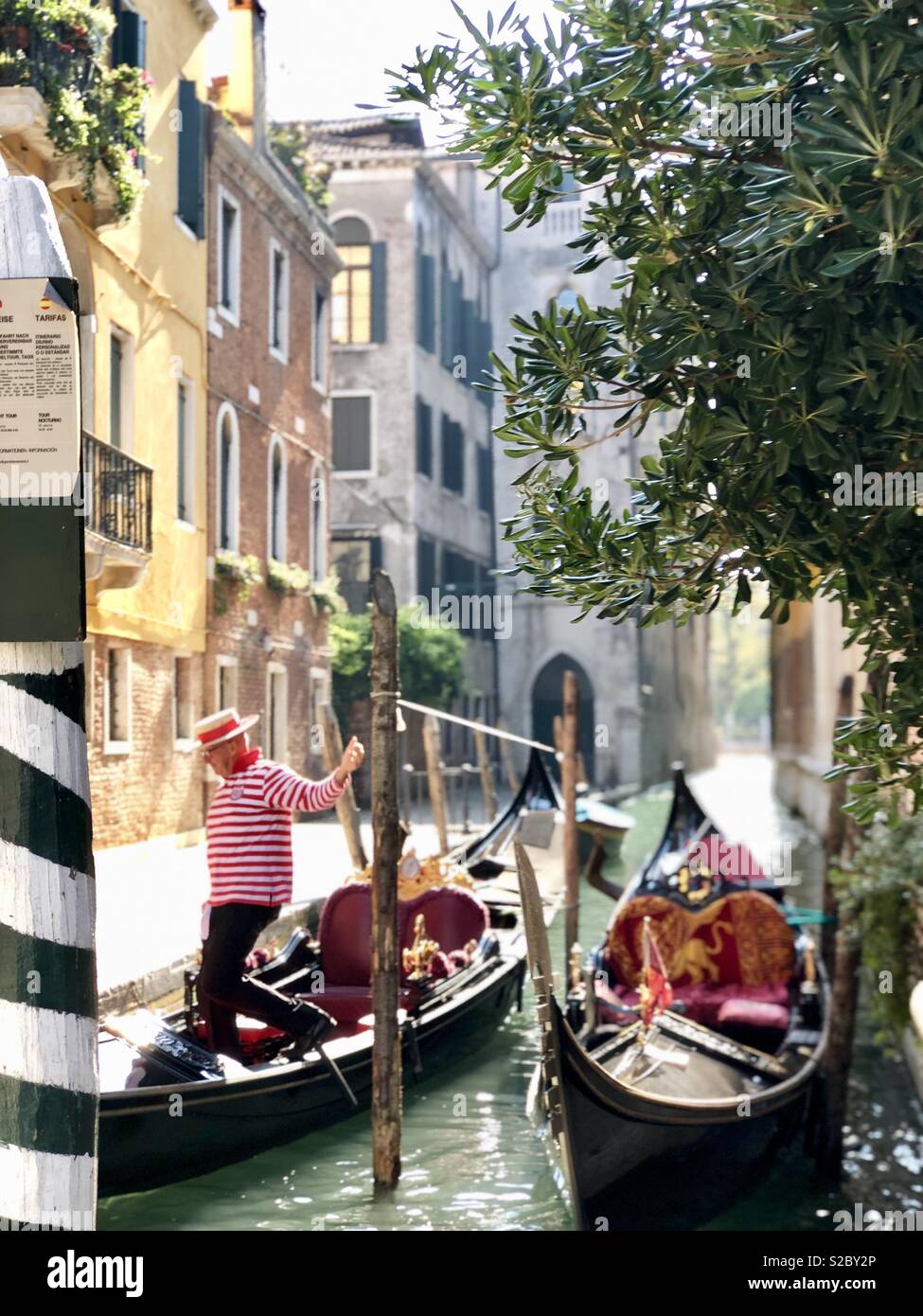Gondola boats and man in red and white striped shirt hopping to shore in small canal in Venice, Italy - Smartphone Captured Stock Image