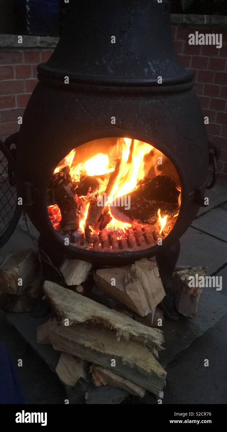 Large black cast iron Chiminea with a fire burning and more wood stacked ready Stock Photo