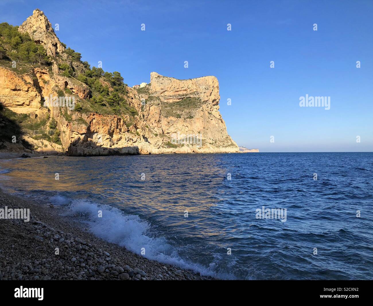 Late afternoon view over pebble beach to dramatic golden sea cliffs, Cala del Moraig, Benitachell, Costa Blanca, Spain Stock Photo
