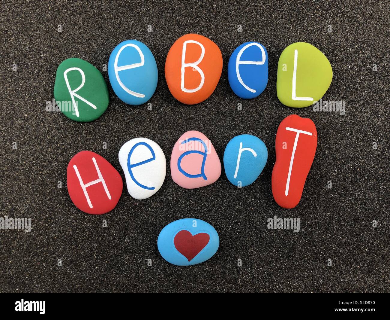 Rebel heart text with colored stones over black volcanic sand Stock Photo