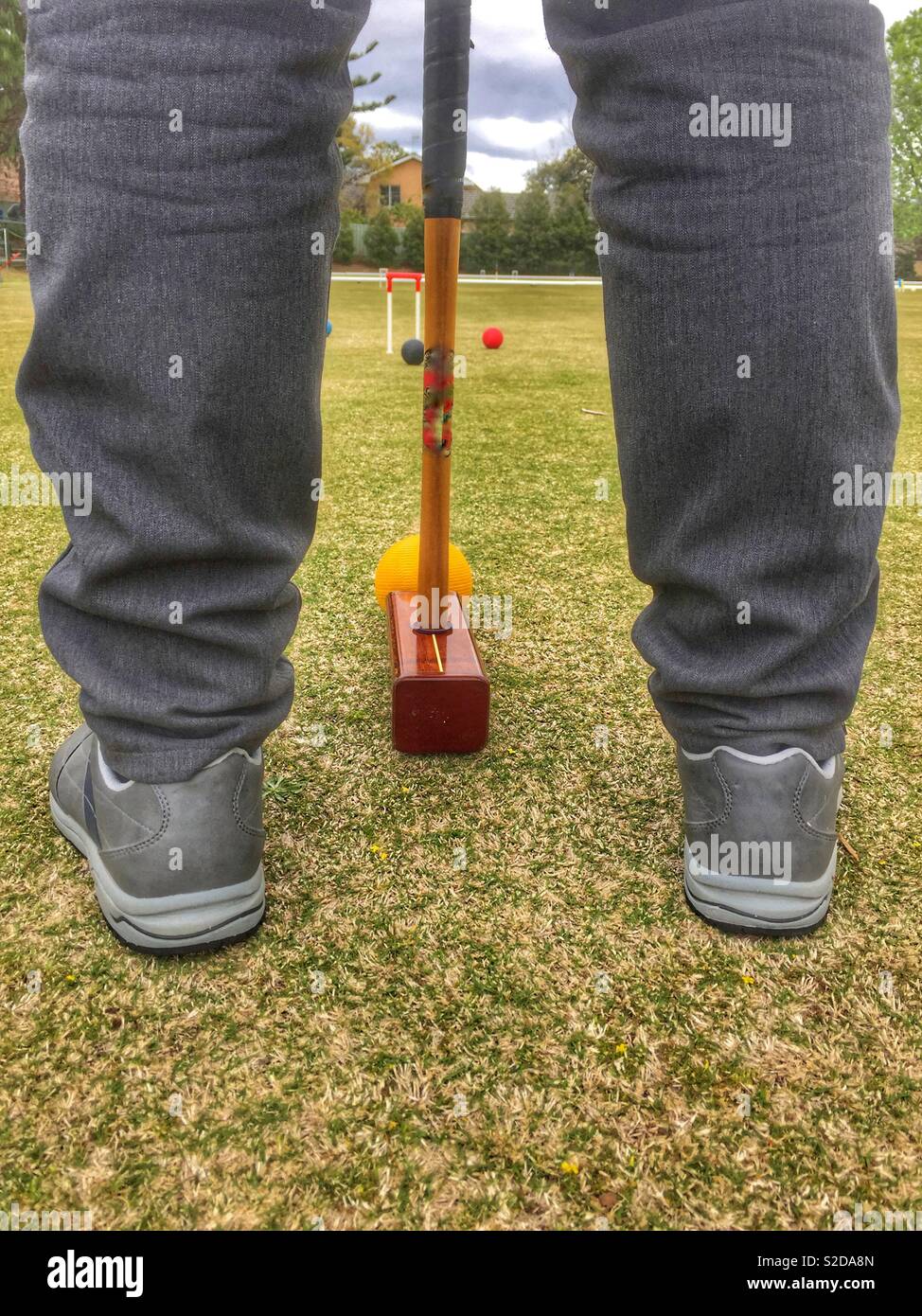 Between the legs rear view of croquet game as player hits ball with mallet. Croquet is  a  strategy / sport played outdoors involving hitting balls with a mallet through hoops on lawn. Stock Photo