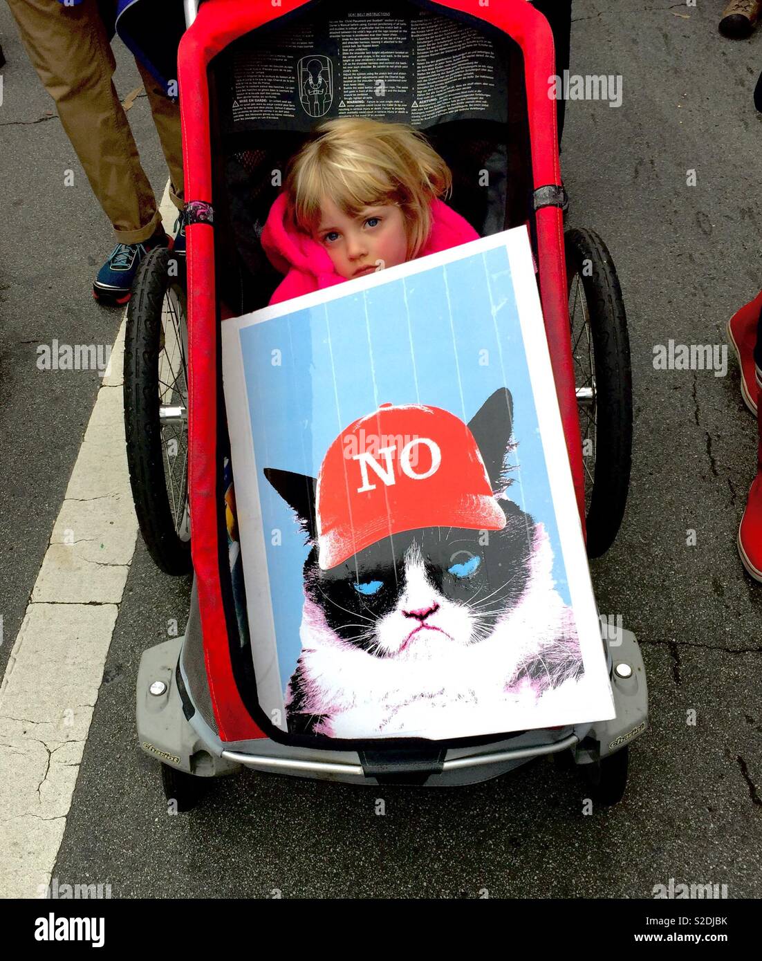Tired blonde child in stroller behind a protest sign of Grumpy Cat wearing a red baseball cap that says NO, instead of a MAGA hat. Women’s March 2017. Oakland, California, USA Stock Photo