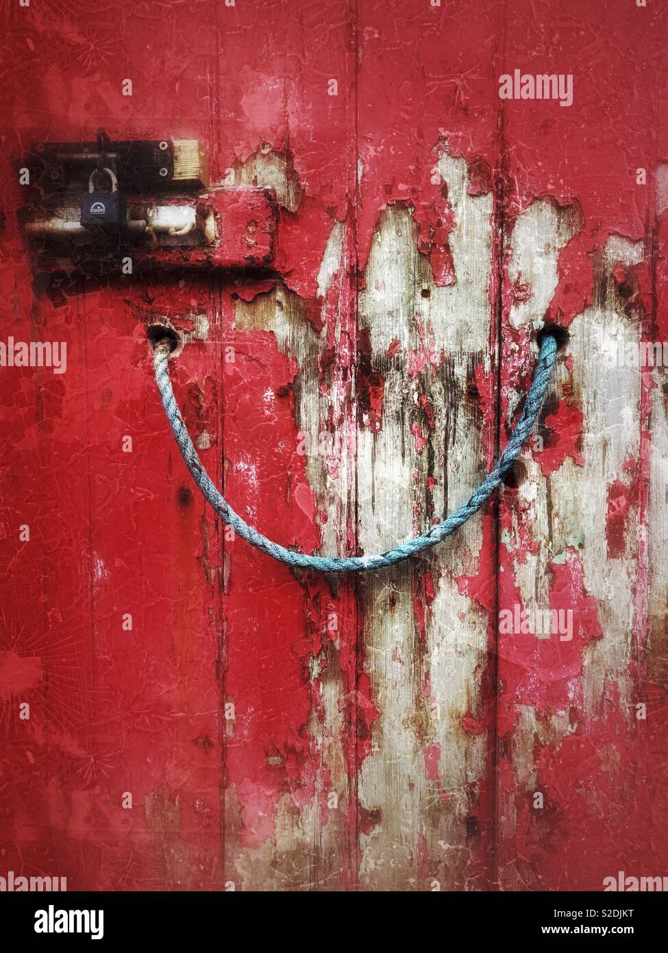Peeling paint on an old red door with blue rope handle and locks. - Smartphone Captured Stock Image