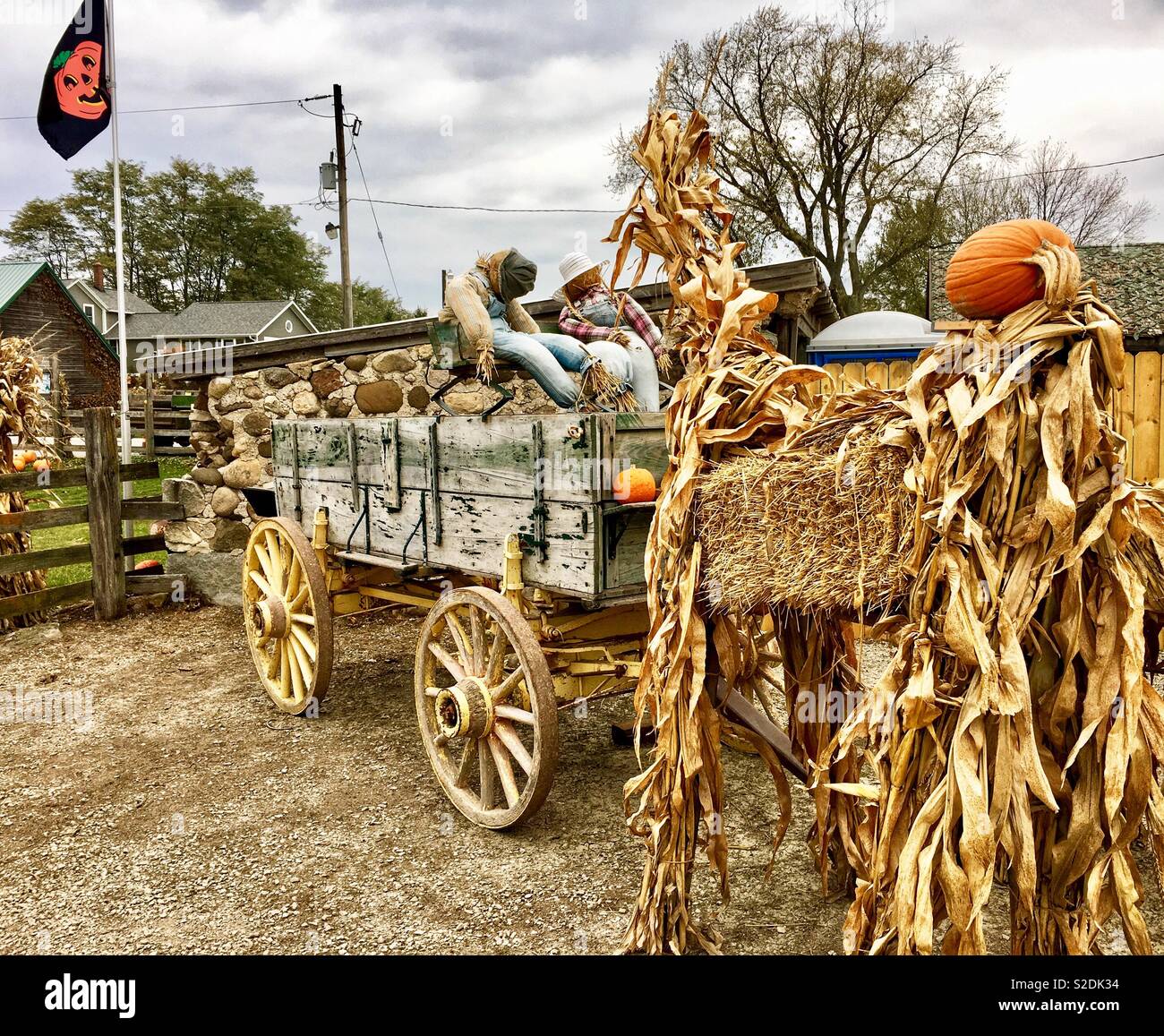Creepy Halloween setup of two scarecrows in overalls driving a wagon with a hay corn pumpkin head monster at the front with Overcast sky and Jack o’ lantern flag - Smartphone Captured Stock Image