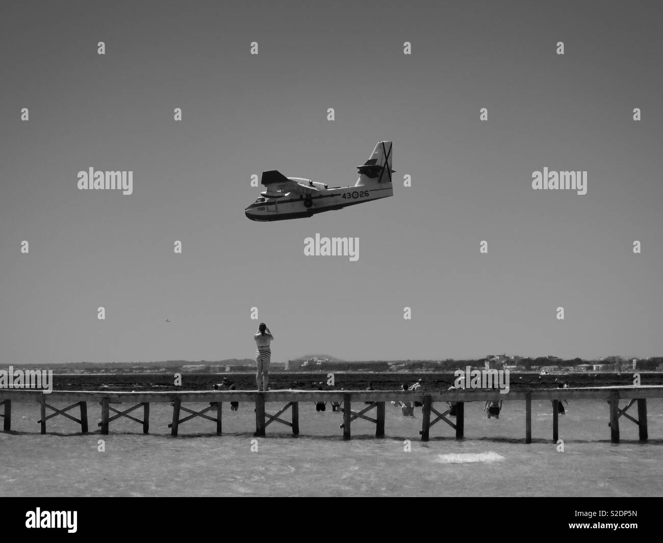 Woman taking a photo of a Fire fighting plane flying past a jetty on a beach in Alcúdia, Majorca, Spain Stock Photo