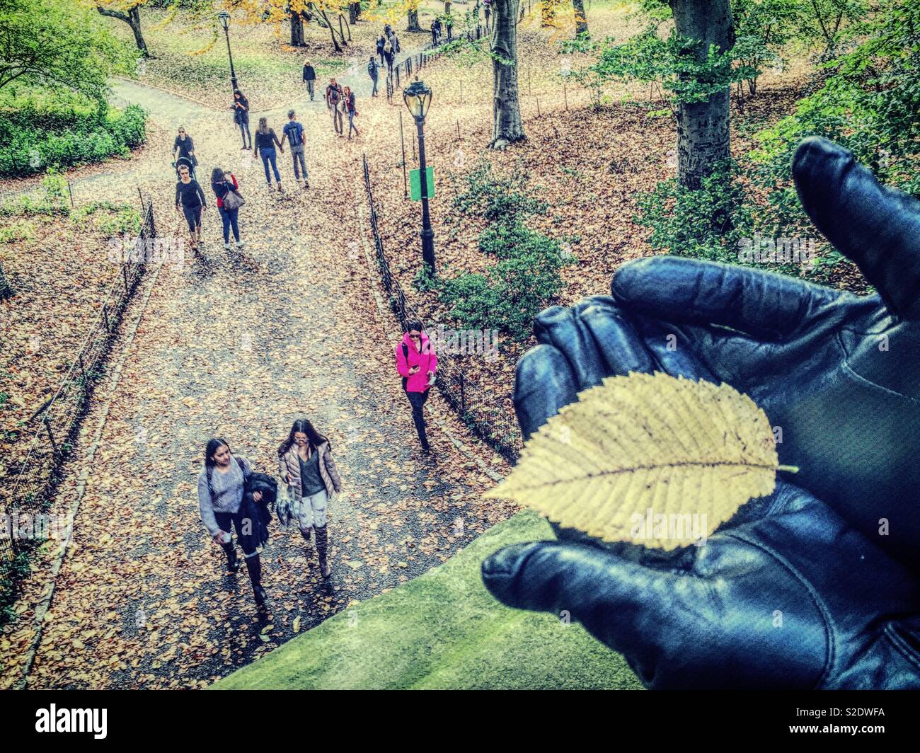 Gloved hand holding a bright fallen leaf as Park goers enjoy the pathway under glade arch near Cedar Hill in Central Park, NYC, USA - Smartphone Captured Stock Image