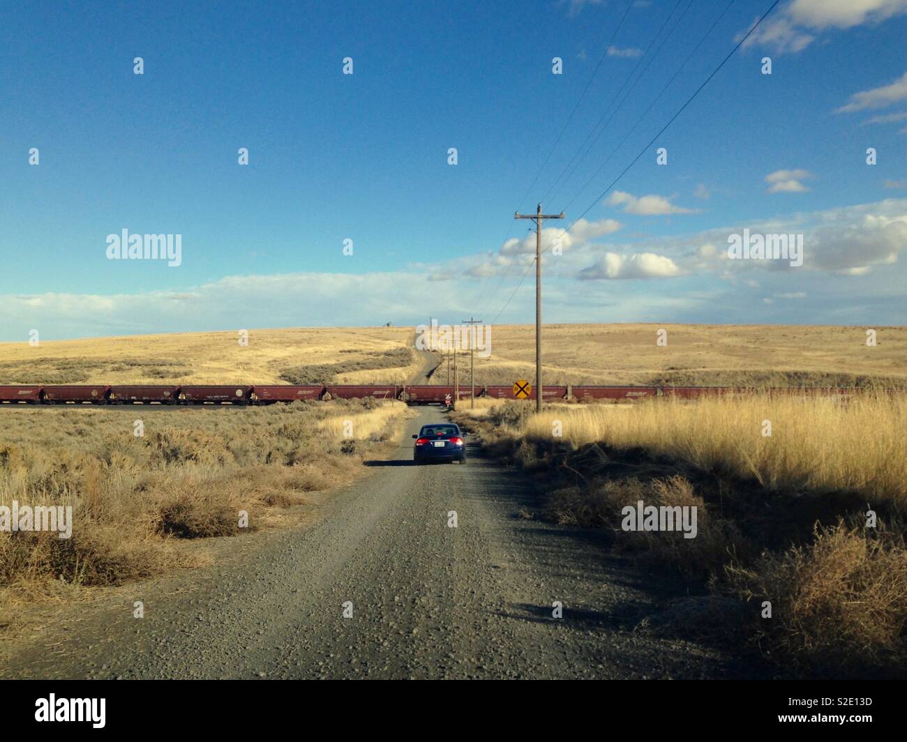 Car stops for a long train on straight gravel road with wheat fields on both sides in southeastern Washington state with wide horizon, blue sky, and expressive clouds Stock Photo