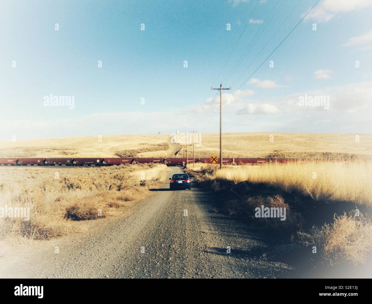 Misty photo of car stopped for a long train on straight gravel road with wheat fields on both sides in southeastern Washington state with wide horizon, blue sky, and expressive clouds - Smartphone Captured Stock Image