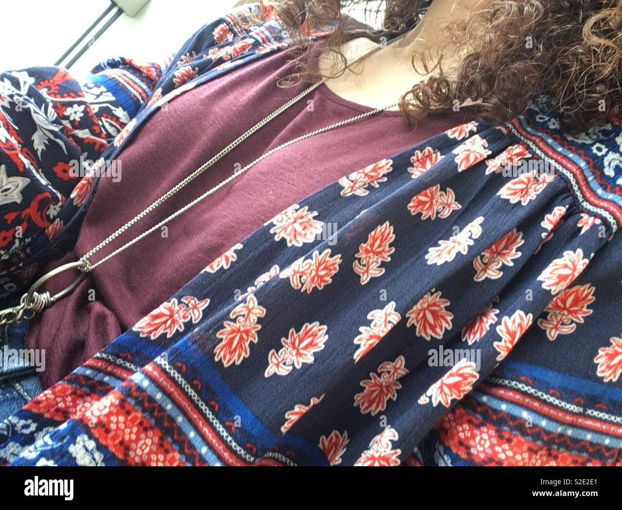 Dark brown curly haired female wearing long silver necklace against plum coloured T-shirt and blue red floral print soft jacket Stock Photo