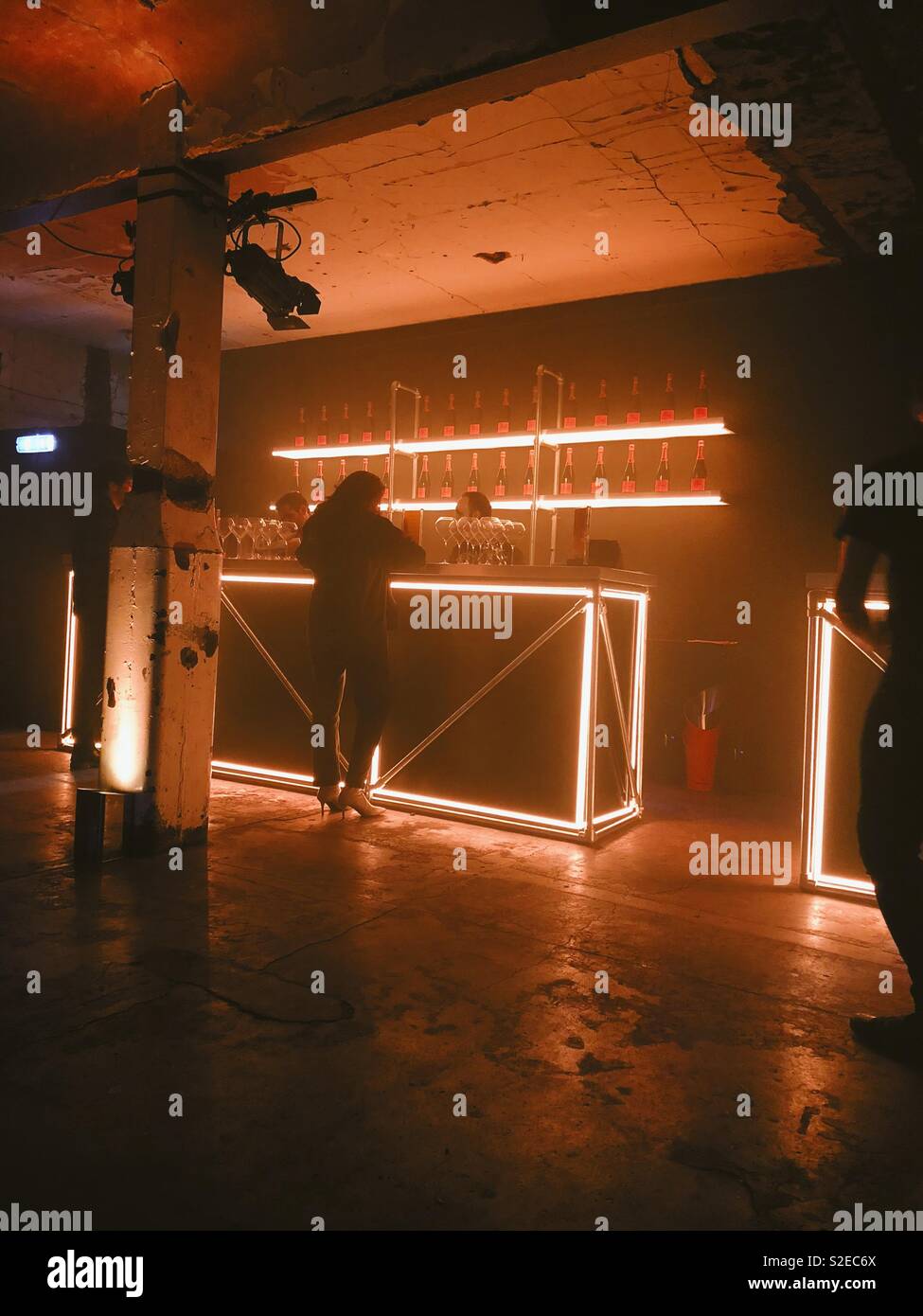 Woman ordering a drink at Champagne bar in warehouse with dramatic lighting and lines of veuve cliquot champagne on the illuminated shelving Stock Photo