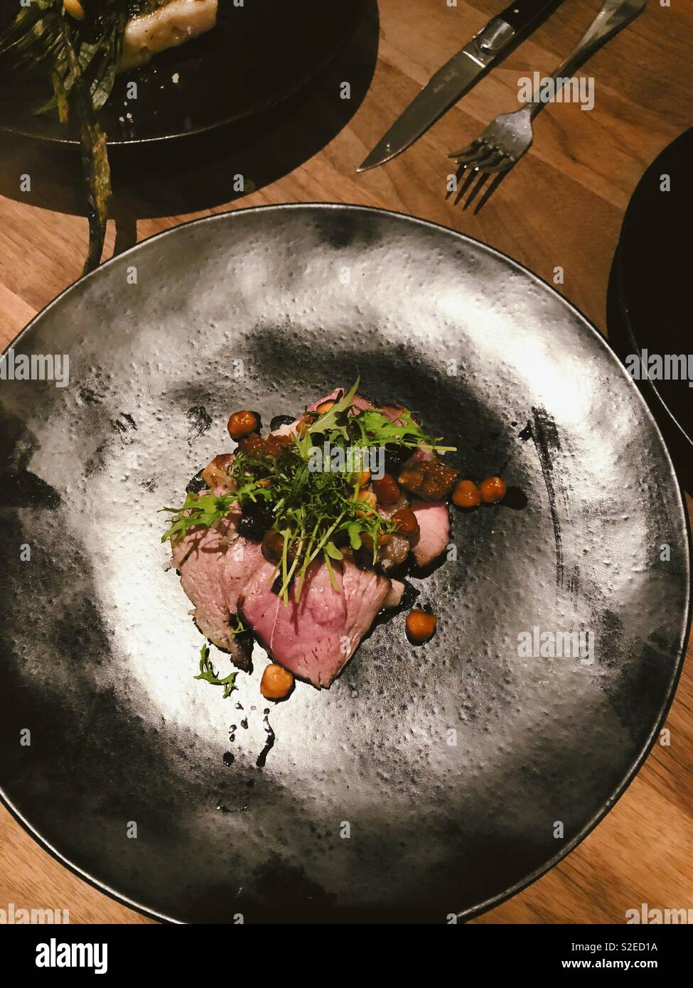 Roast lamb with hazelnuts and leaf garnish on black matte plate on wooden table Stock Photo