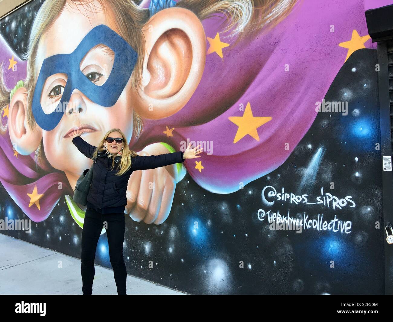 Attractive, young blonde woman, tour operator, with arms outstretched in front of street art by the Bushwick Collective, in Brooklyn, New York - Smartphone Captured Stock Image