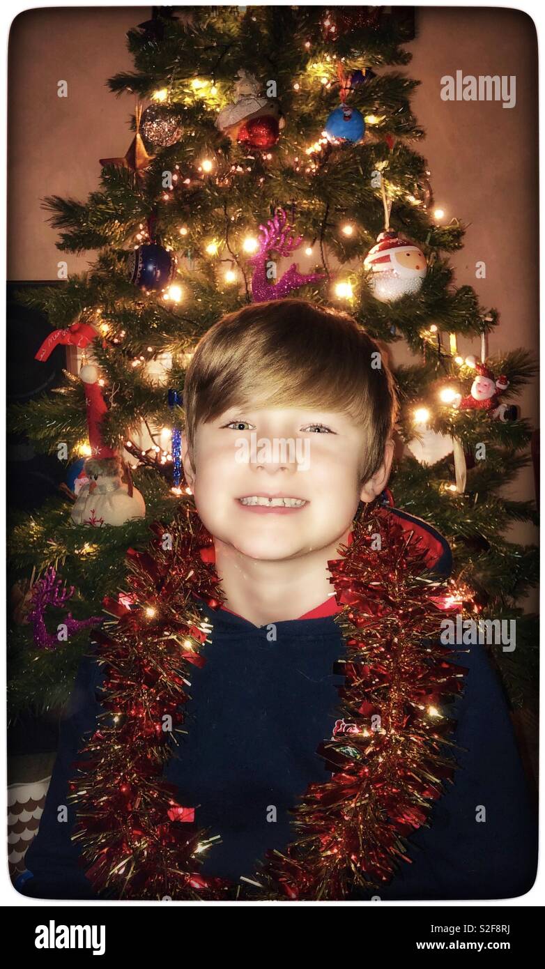 Young boy decorated in tinsel in front of a Christmas Tree. Stock Photo