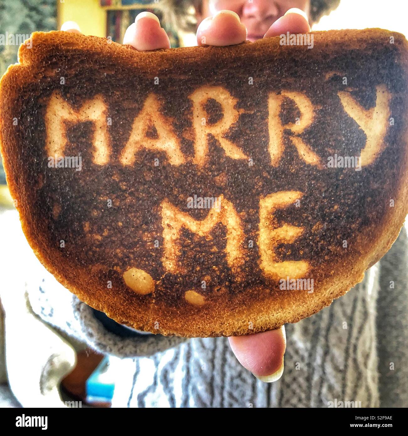 Creative love and romance concept.  Woman holding up a piece of burnt toast with the words Marry Me scraped on the surface. - Smartphone Captured Stock Image