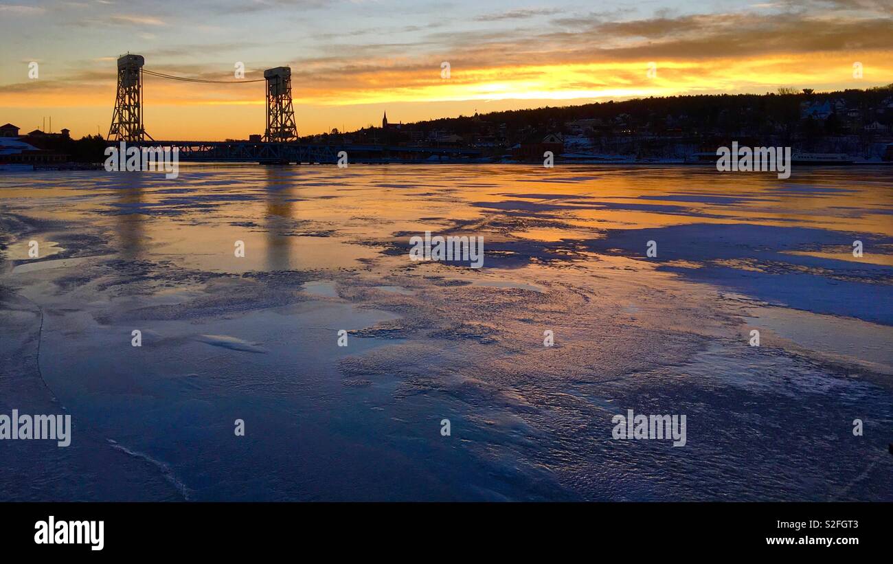 Frozen river at sunrise in Houghton, Michigan, in December with buildings silhouetted and sky reflected in ice Stock Photo