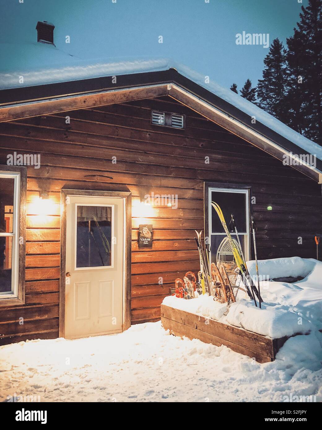 Several sizes of snowshoes and skis outside the door of a house at dusk.  It looks cozy inside after a day enjoying the snow. - Smartphone Captured Stock Image