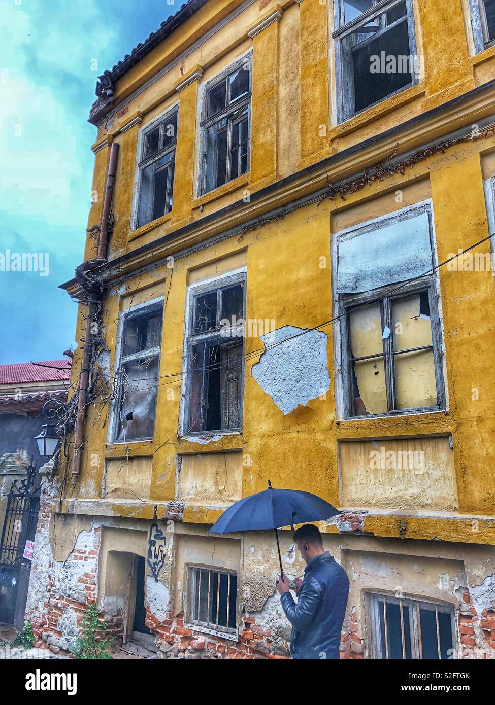 A man holding a black umbrella stands in front of an old run down historical building in the Old Town of Plovdiv, Bulgaria. - Smartphone Captured Stock Image