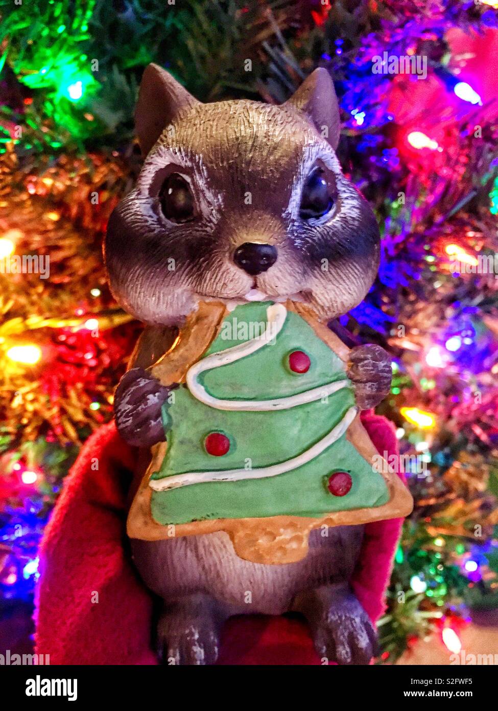 Christmas lawn ornament of chipmunk eating Christmas cookie in front of lit Christmas tree with colorful lights held by red gloved hand - Smartphone Captured Stock Image