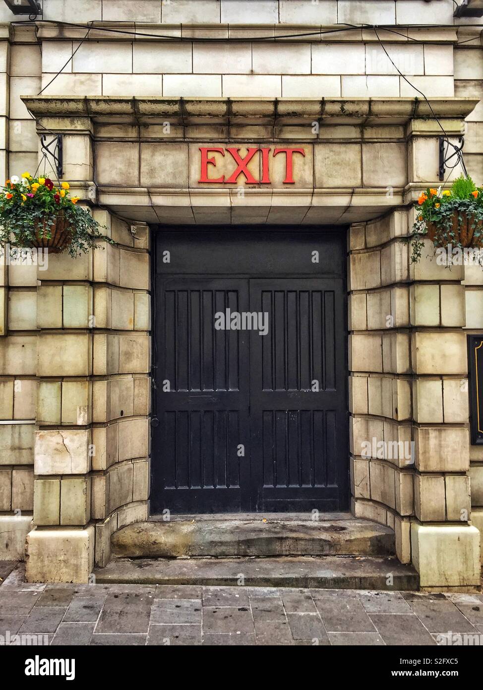 A bright red exit sign above an austere-looking door set in heavy stone wall of the Coliseum, a Witherspoons pub in Abergavenny, Wales, UK. - Smartphone Captured Stock Image