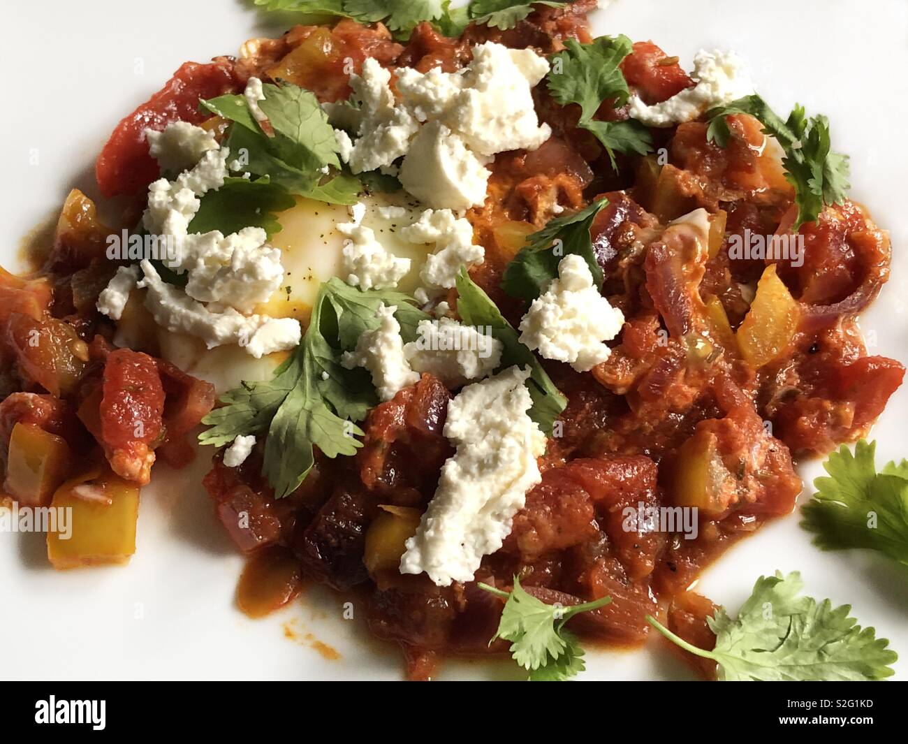 A single serving of the middle eastern dish shakshuka or shakshouka, a poached egg cooked with tomatoes, onions and peppers and topped with feta cheese and cilantro leaves. - Smartphone Captured Stock Image
