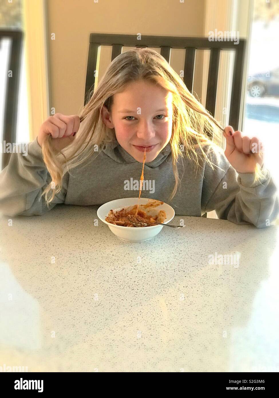 A girl holds her long hair as she slurps spaghetti from a bowl. - Smartphone Captured Stock Image