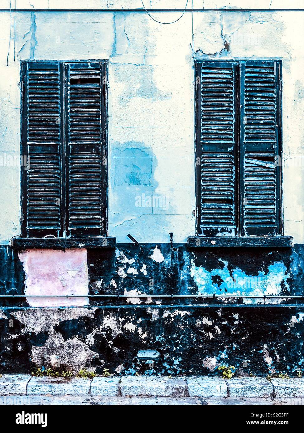 Broken wooden shutters with peeling paint on windows and wall below Stock Photo