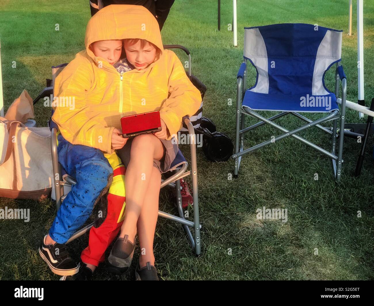 Two children share a jacket and a game while waiting at an outdoor event. - Smartphone Captured Stock Image