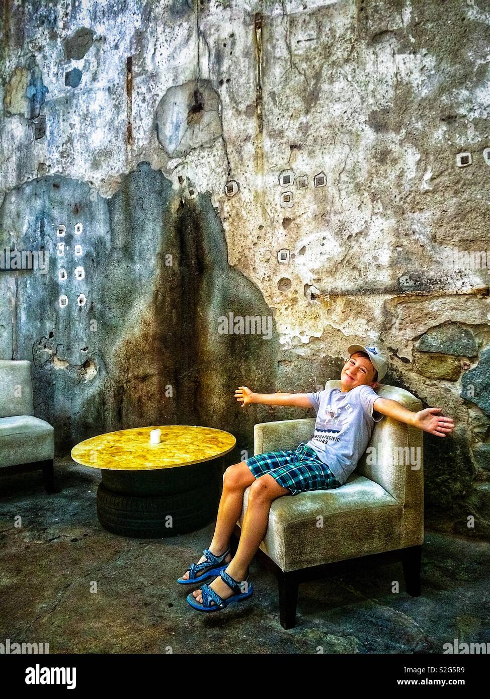 Smiling boy seated on an vintage armchair in the hall of an artistic and old industtial building - Smartphone Captured Stock Image