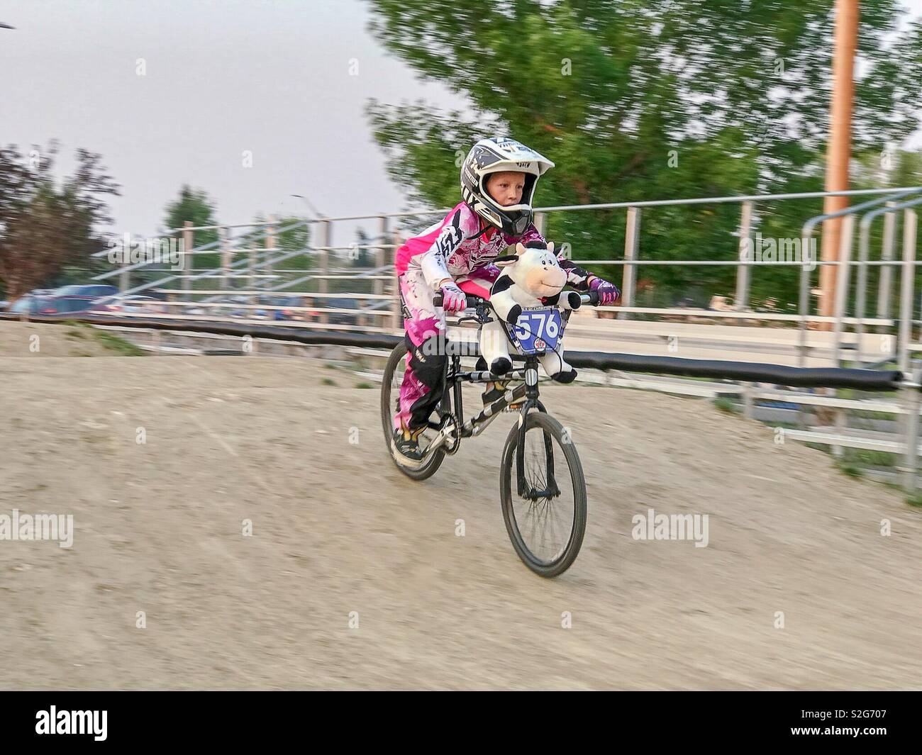 A girl races on a BMX track with her toy strapped to the handlebars. Stock Photo