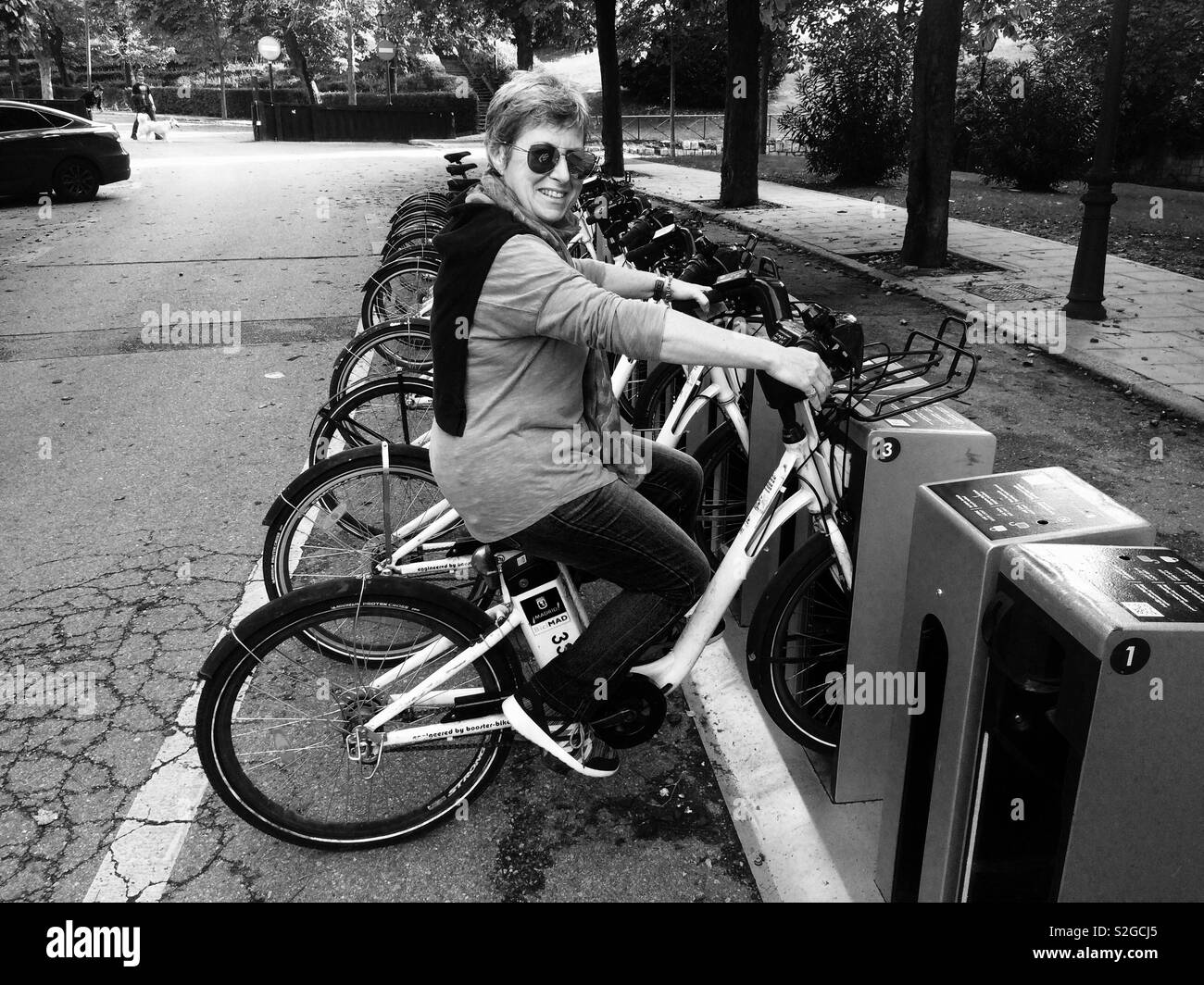 Mature woman wearing sunglasses and smiling sitting on electric bicycles in their docking station in Madrid Spain Stock Photo