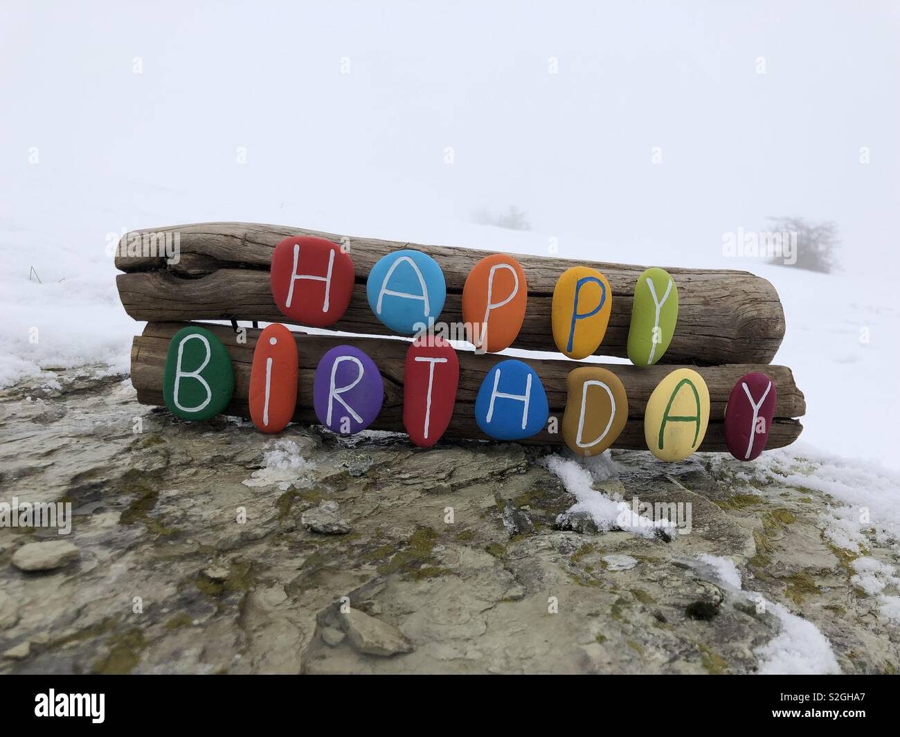 Happy Birthday message with a colored stones composition over woods and snow background - Smartphone Captured Stock Image