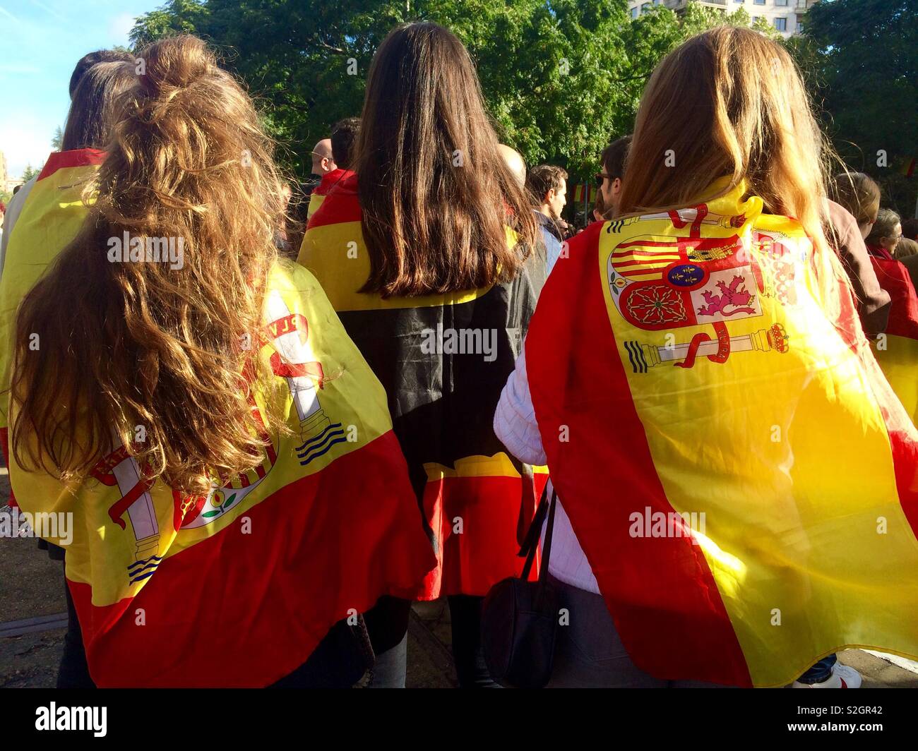 Spanish flags wrapped around the shoulders of three women watching the national day celebrations in the streets of Madrid in Spain Stock Photo