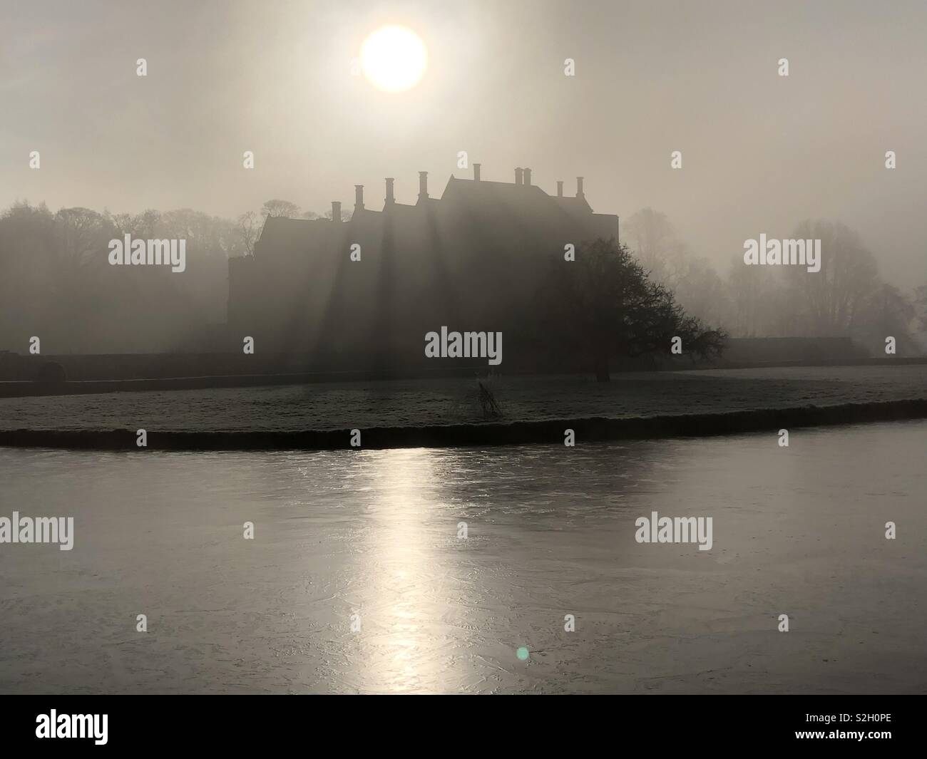 Broughton Castle framed by its frozen moat during a hard frost and dramatically silhouetted by the rising Sun with crepuscular rays created by the tall Tudor chimneys. - Smartphone Captured Stock Image
